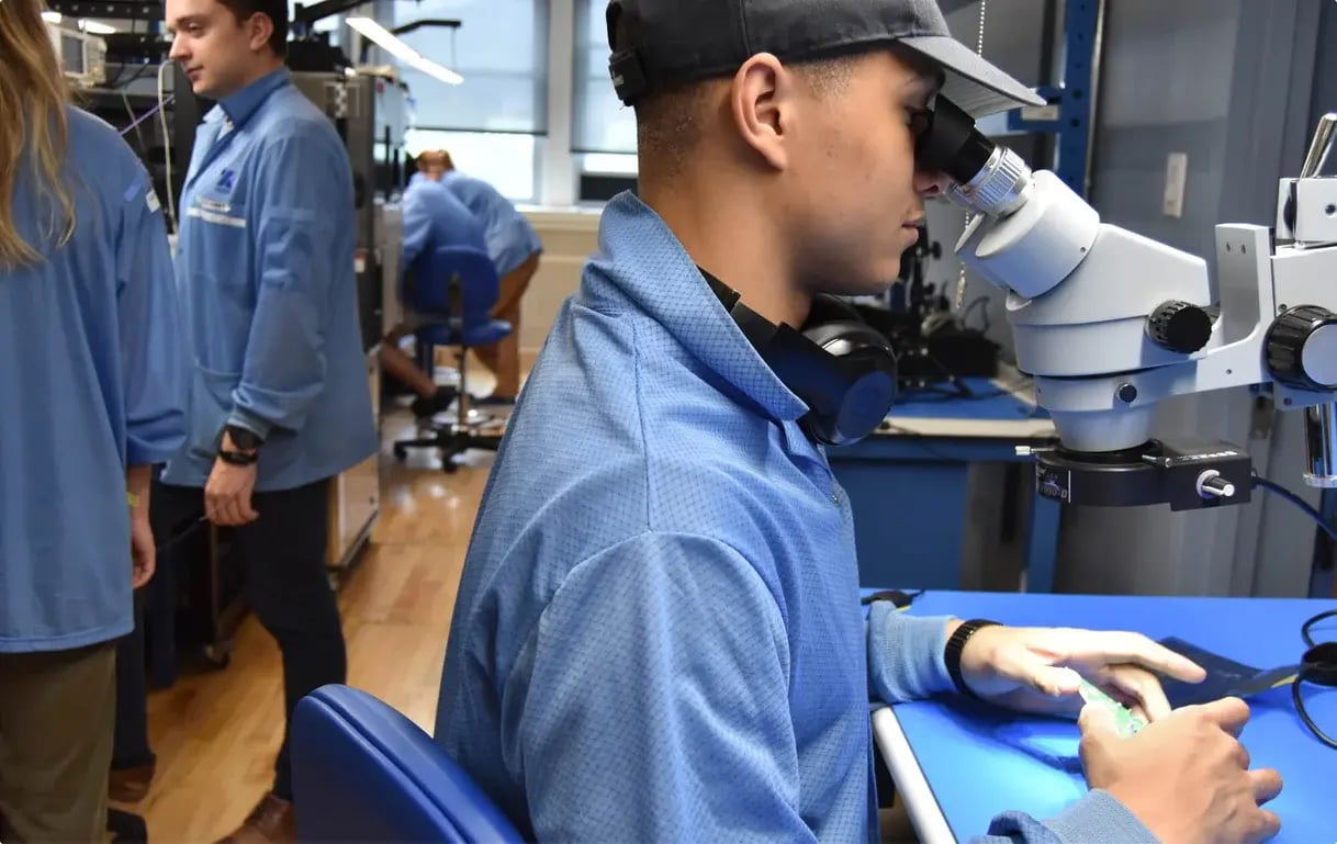 A person wearing a blue lab coat and a baseball cap looks through a microscope at a circuit board on a blue desk in a lab setting, with other people in the background.
