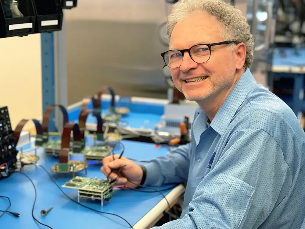 A smiling man in glasses and a blue shirt works at a workbench, testing electronic components. He's holding a tool, focused on a circuit board.